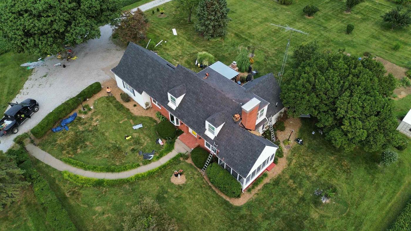 Two crew members working on a shingle roof of a large brick farmhouse, seen from above, with a ladder against the house and materials staged on the driveway